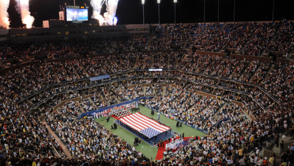 Nighttime aerial view of Arthur Ashe Stadium during the US Open, featuring a large American flag display and a cheering crowd under bright stadium lights.