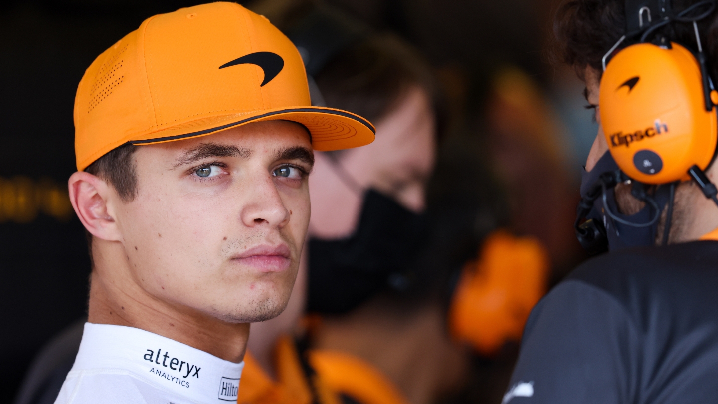 Formula 1 driver in an orange team cap looking focused in the paddock, surrounded by the pit crew.