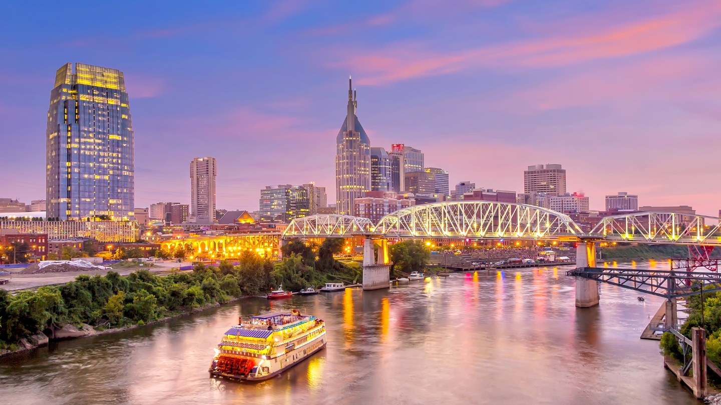 The Nashville city skyline illuminated at sunset with the iconic bridge and a cruise boat on the river in the foreground.