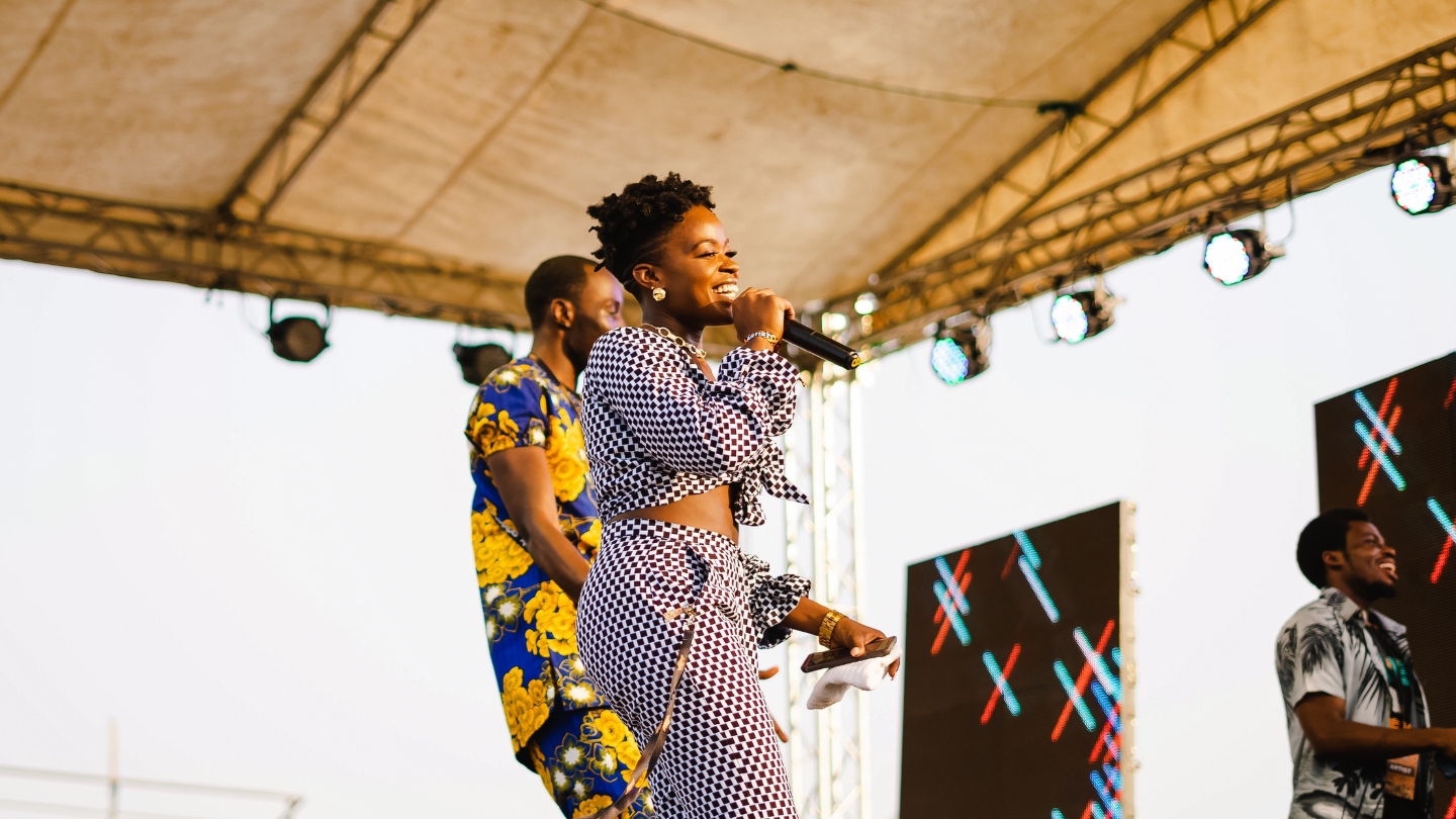 A smiling female artist performing on an outdoor stage, engaging the audience during a vibrant live music festival.