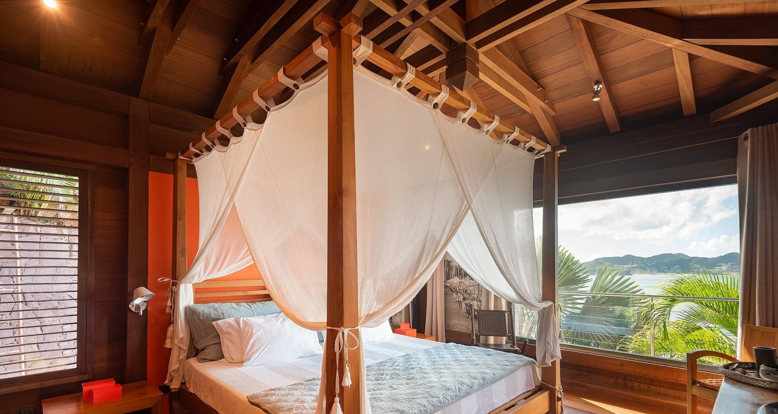 Canopy Bed with Ocean View – A cozy bedroom retreat with a luxurious canopy bed, wooden accents, and floor-to-ceiling windows overlooking the turquoise waters of St. Barths.