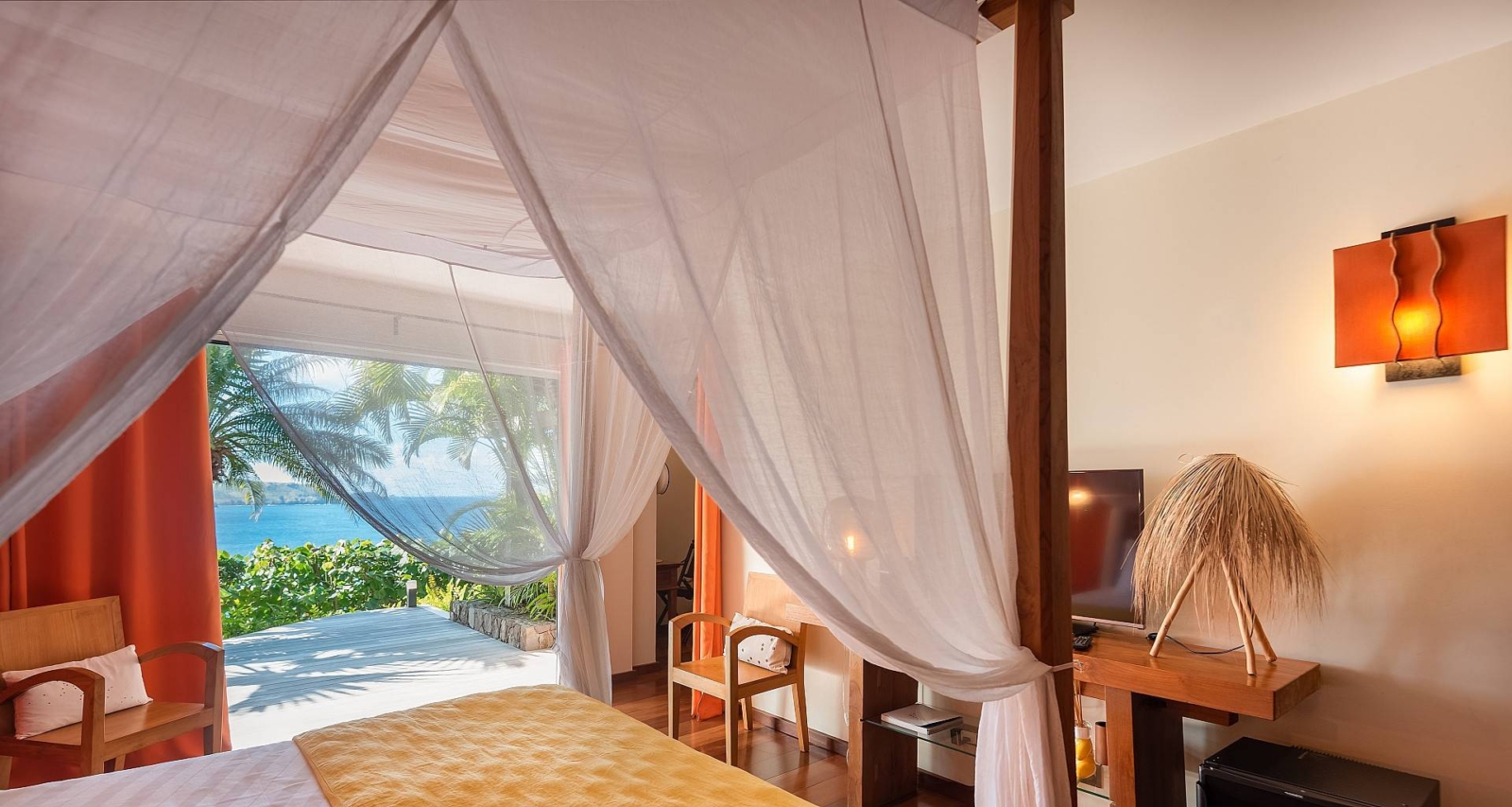 Cozy bedroom with soft lighting, wooden accents, and a desk area, featuring an ocean view through the open-air terrace.