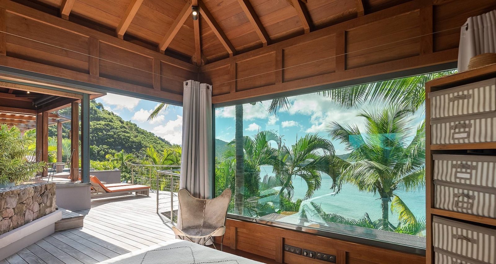 Spacious bedroom with a floor-to-ceiling glass window offering an immersive ocean view, surrounded by tropical palm trees.