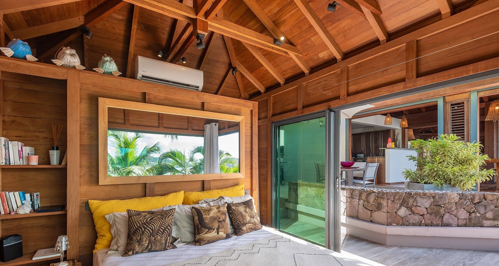 Open-concept bedroom with tropical wood elements, a cozy bed with vibrant pillows, and a large mirror reflecting the lush outdoor scenery.