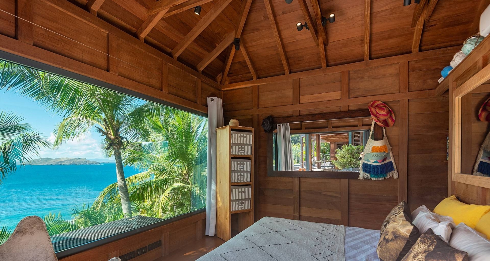 Scenic bedroom with a panoramic ocean view framed by palm trees, a stylish bookshelf, and natural wood accents.