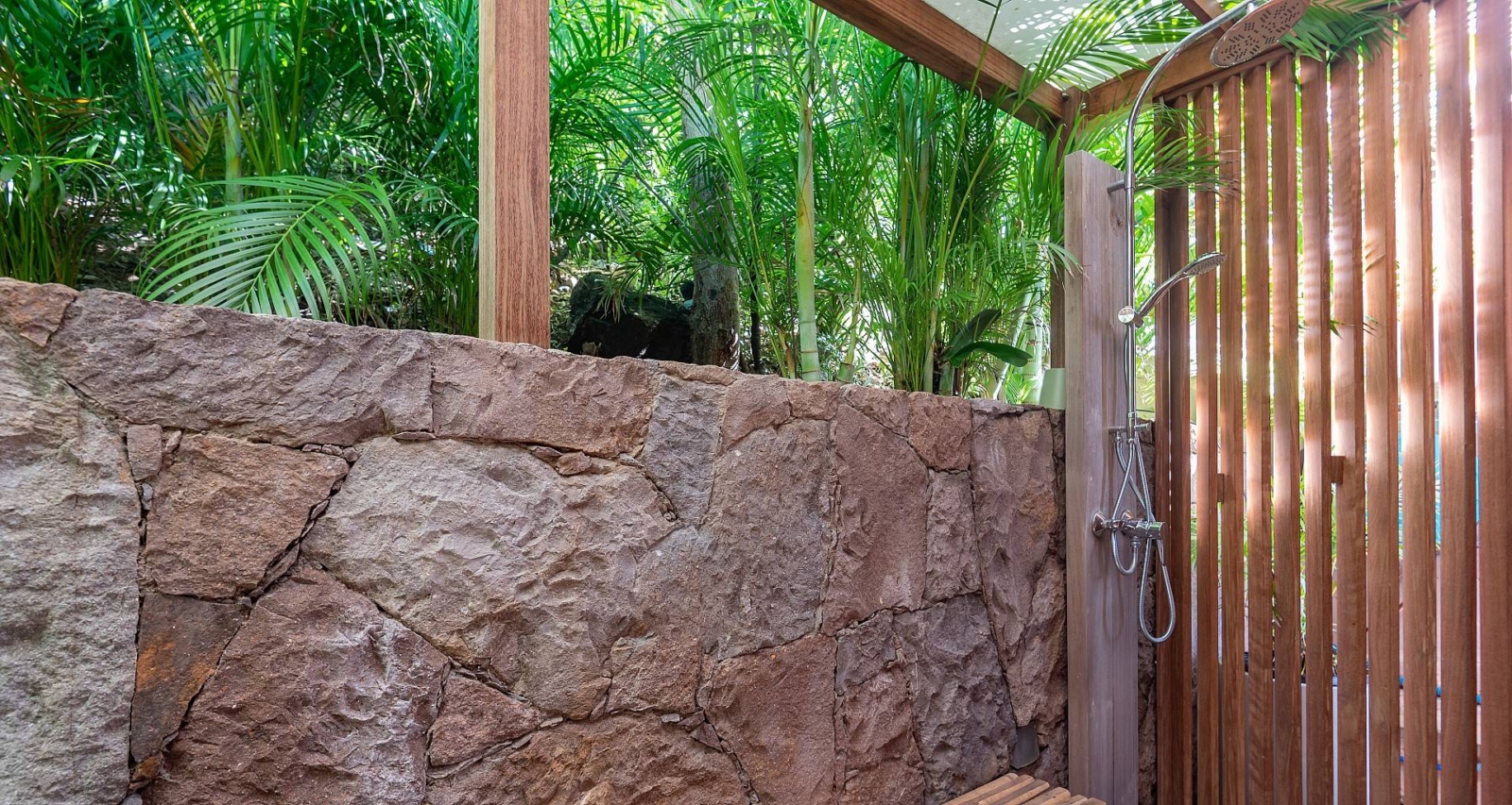 Serene outdoor shower with a stone wall, wooden slats, and surrounded by tropical plants, creating a relaxing, nature-inspired retreat.