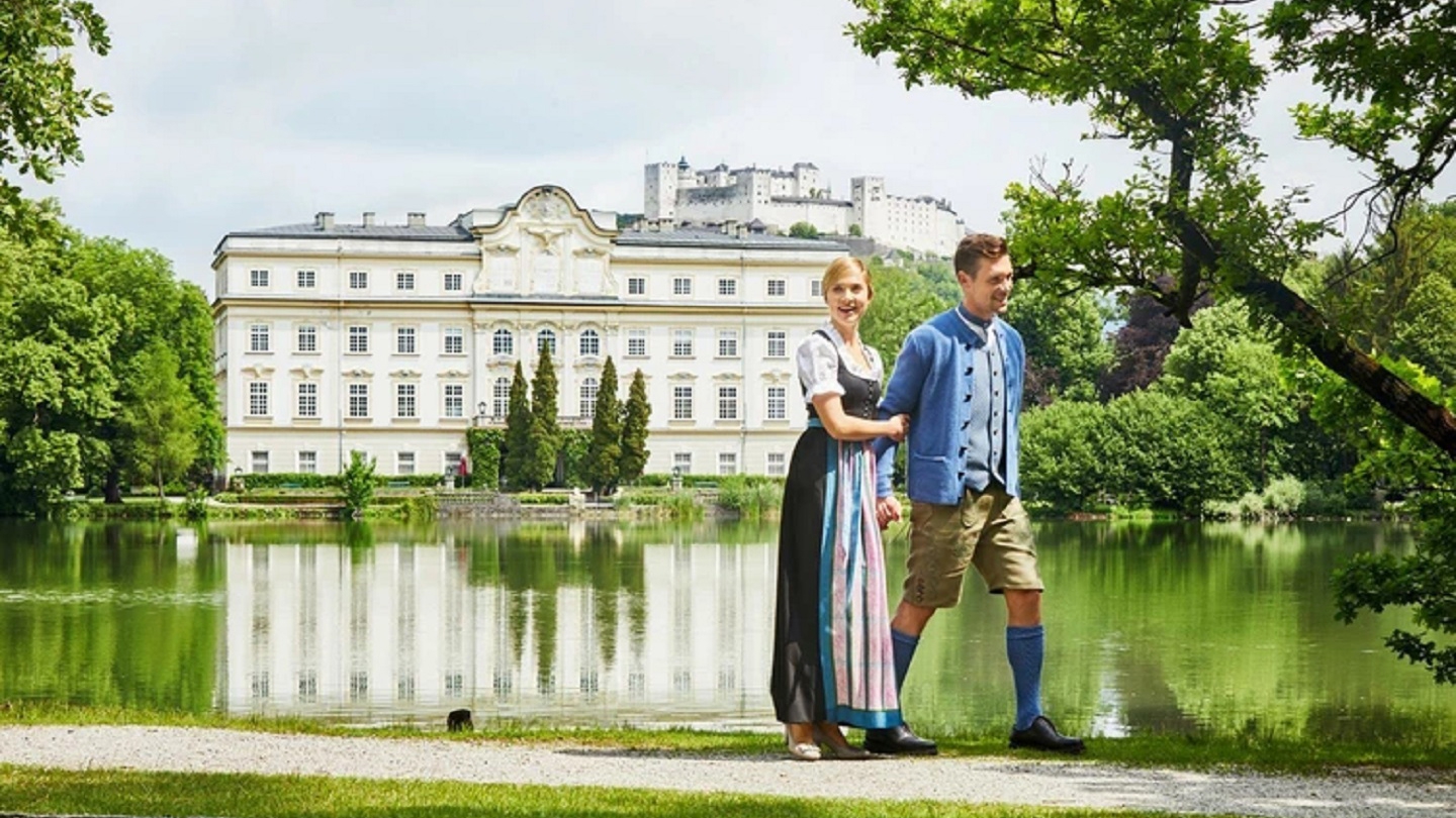 Charming couple dressed in traditional Austrian attire walking near the historic Leopoldskron Palace in Salzburg.