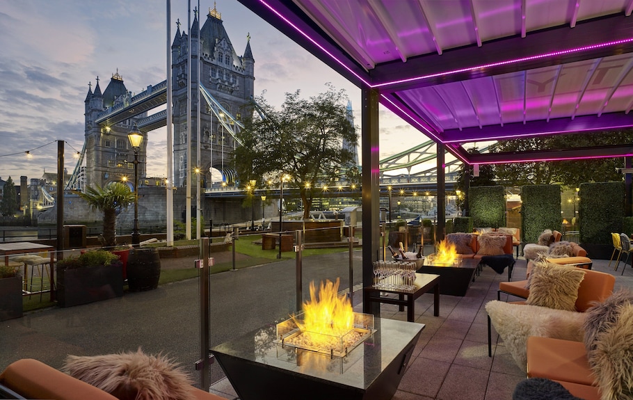 Scenic view of Tower Bridge from a stylish riverside lounge in London, featuring cozy outdoor fire pits and elegant seating.