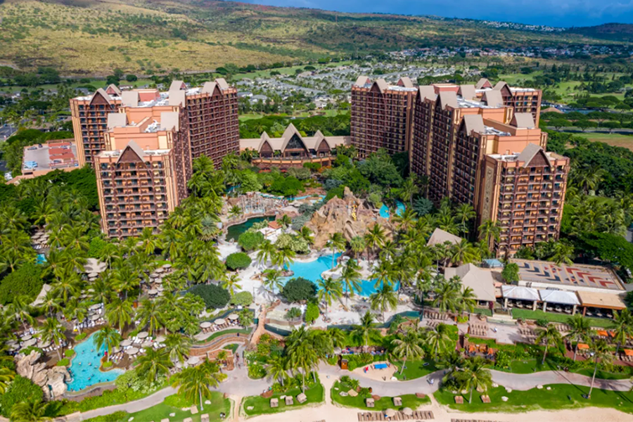 A breathtaking aerial perspective of Aulani, a Disney Resort & Spa, showcasing its lush greenery, towering accommodations, and vibrant pool areas set against a backdrop of rolling hills.