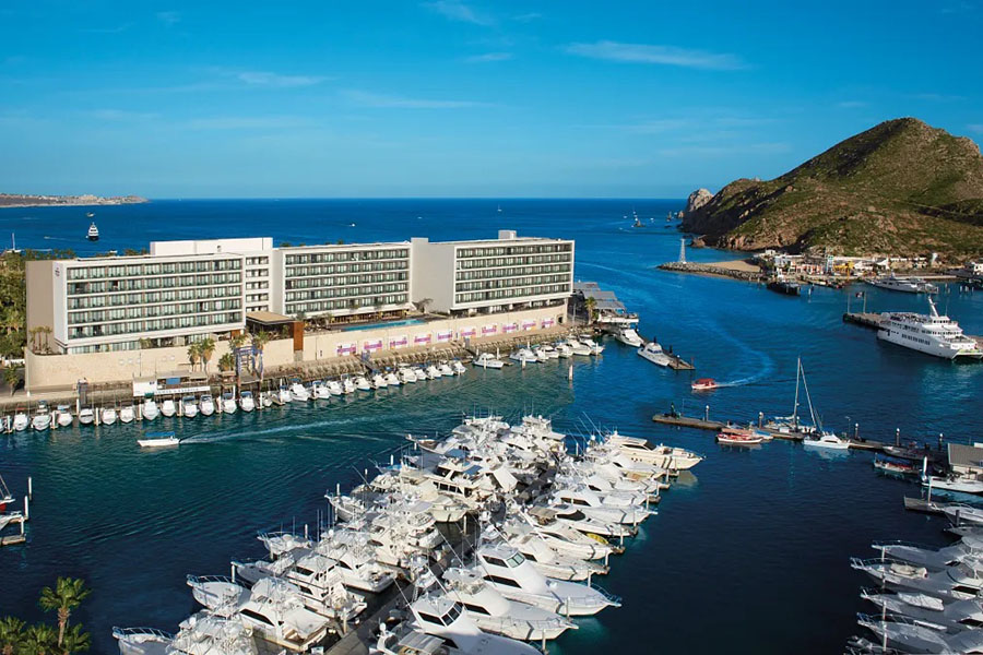 A stunning view of the Breathless Cabo San Lucas Resort & Spa overlooking a luxurious marina filled with yachts, set against the backdrop of clear blue waters and rolling hills.