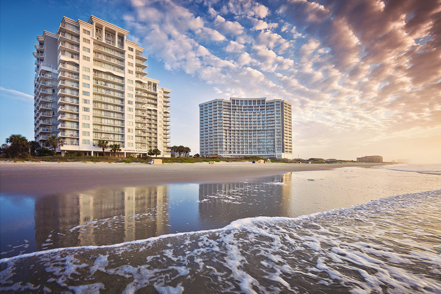 Elegant beachfront resort with towering modern buildings reflecting the golden hues of sunrise, set against a backdrop of soft waves and a dramatic sky, offering a perfect coastal retreat.