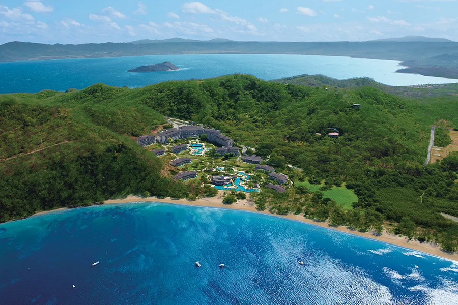 A spectacular aerial view of the Dreams Las Mareas Costa Rica, nestled along a pristine bay with lush tropical mountains in the background.