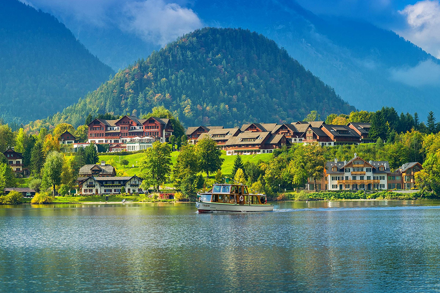 Charming lakeside resort surrounded by lush greenery and nestled at the base of a picturesque mountain, featuring traditional alpine architecture and a serene boat gliding across the water.