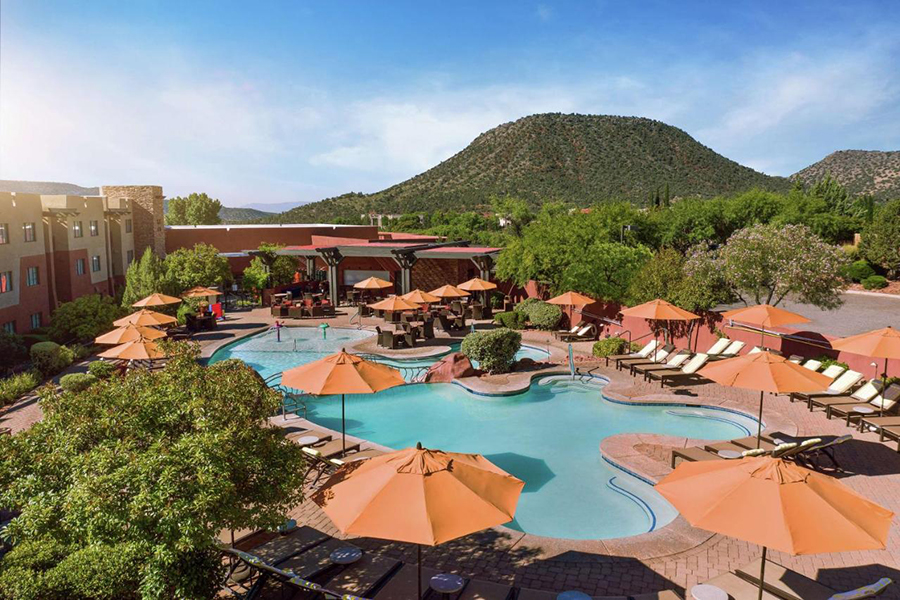 Relaxing resort featuring a sparkling outdoor pool surrounded by orange umbrellas and lush greenery, set against the stunning backdrop of Sedona's iconic red rock mountains.