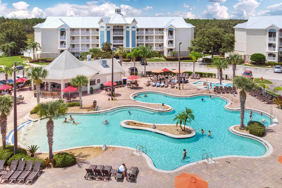 Family-friendly resort featuring a large, winding pool surrounded by palm trees, lounge chairs, and vibrant umbrellas, set against charming white buildings and sunny blue skies for the ultimate tropical getaway.