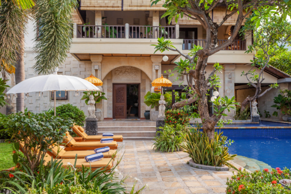 The elegant entrance of a Balinese villa, featuring a pool surrounded by sun loungers, tropical plants, and traditional decor.